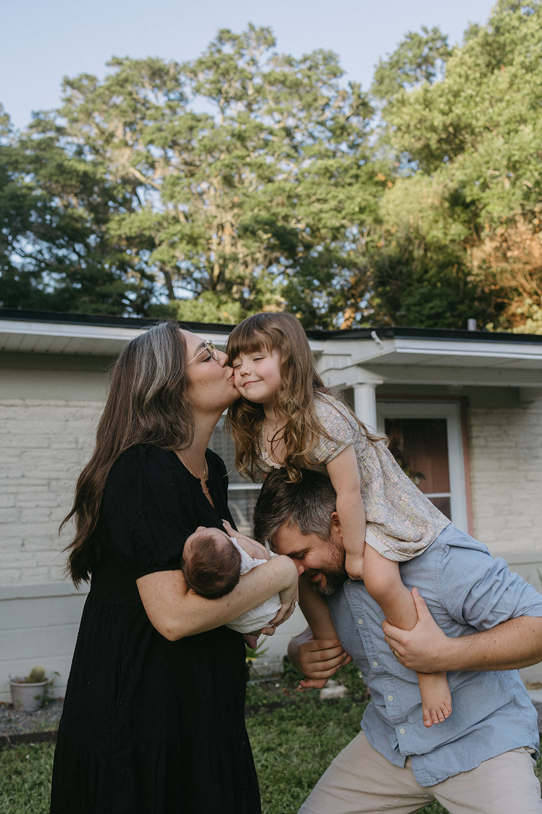 Family spending time outside together. Dad holding sibling on shoulders while mom holds newborn in arms. Mom gives kiss to daughter on the cheek.