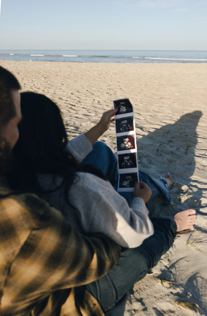 Pregnancy announcement photos in Jacksonville, Fl. Couple sitting on the beach viewing strip of ultrasound pictures.